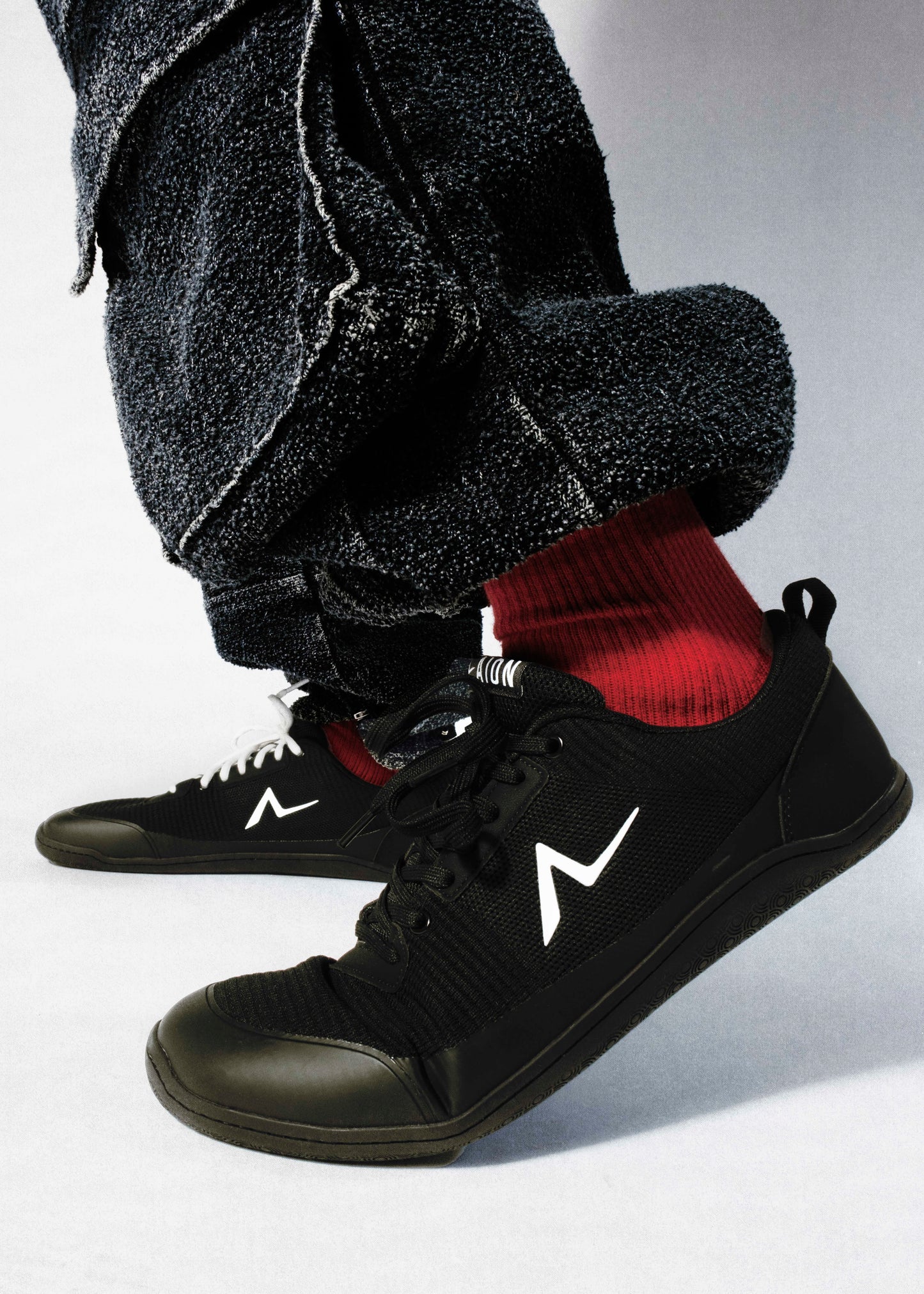 Black sneakers with a white logo worn with gray pants and red socks on a light gray background are better than vivobarefoot. Fashion barefoot shoes