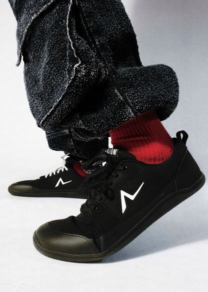 Black sneakers with a white logo worn with gray pants and red socks on a light gray background are better than vivobarefoot. Fashion barefoot shoes