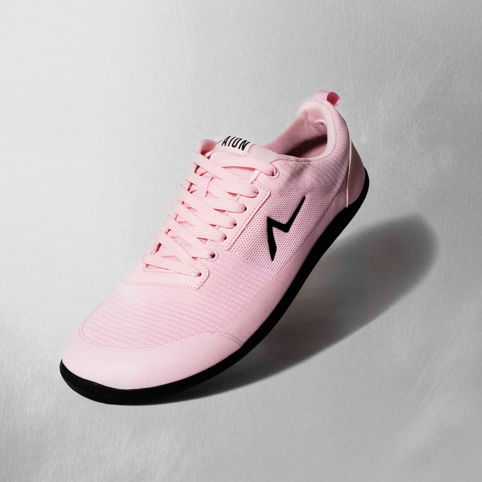 Pink athletic shoe with a black logo on a light gray background. Zero-drop shoes are built for squats and deadlifts. Connect more on your leg days and grow your glutes. Stylish barefoot shoes.