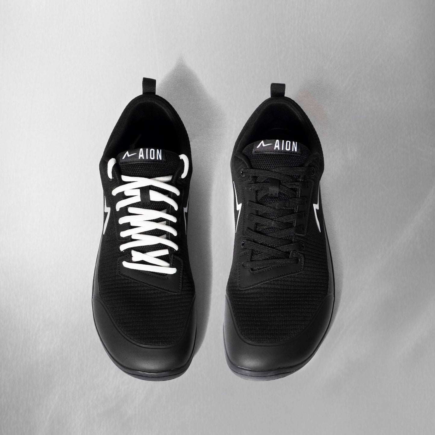 Black athletic shoes with white laces on a light gray background. The best barefoot shoe in the game. Wide toe box flexible sole and the durability to last a long time under strain and load.